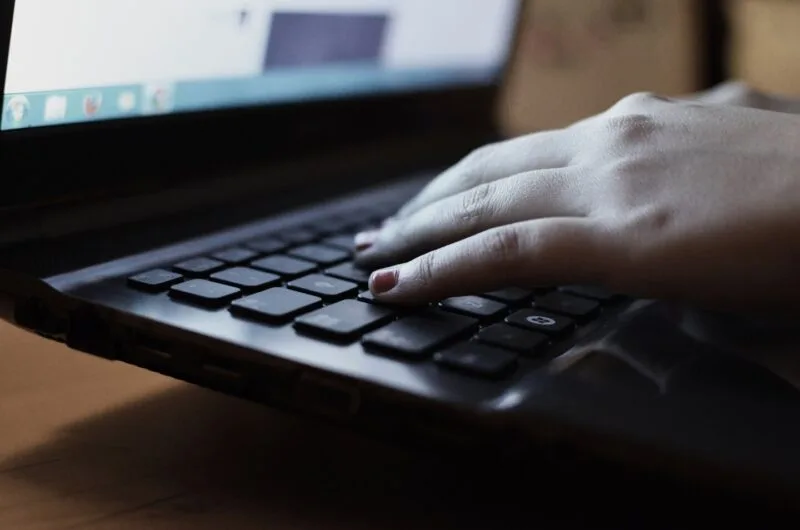 Mâini care tastează pe un laptop, lângă un caiet și un pix