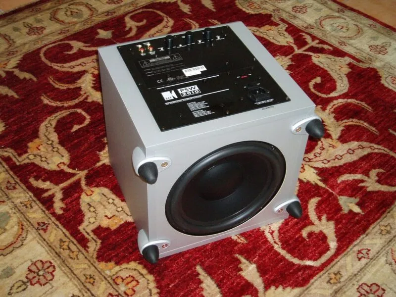 Subwoofer negru KEF PSW2010 așezat pe parchet, lângă perete