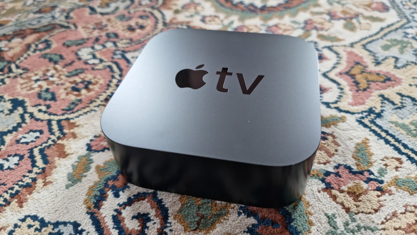 Cutia neagră Apple TV 4K, cu logoul Apple TV pe capac, așezată pe un covor cu model floral