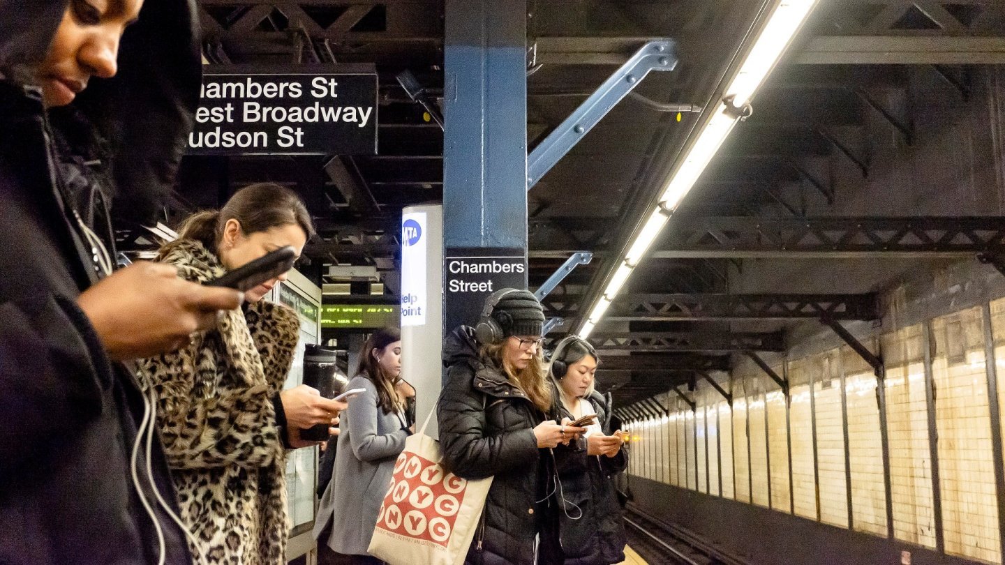 Călători pe peronul unei stații de metrou din New York, cu privirea în ecranele smartphone-urilor
