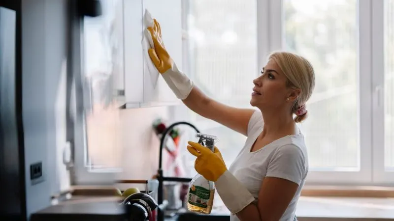 Femeie care curăță dulapurile din bucătărie cu un spray de curățat și mănuși de protecție