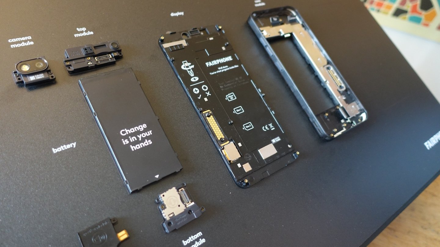 Componentele unui Fairphone 3 expuse separat pe o placă neagră: baterie, cameră, difuzor, ecran și carcasă