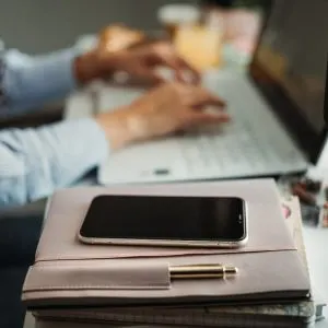 Femeie care tastează la un laptop pe un birou, cu un smartphone și o agendă roz în prim-plan
