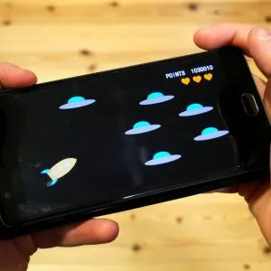 Două mâini țin orizontal un smartphone pe care rulează un joc 2D simplu cu o rachetă și farfurii zburătoare, deasupra unei mese de lemn