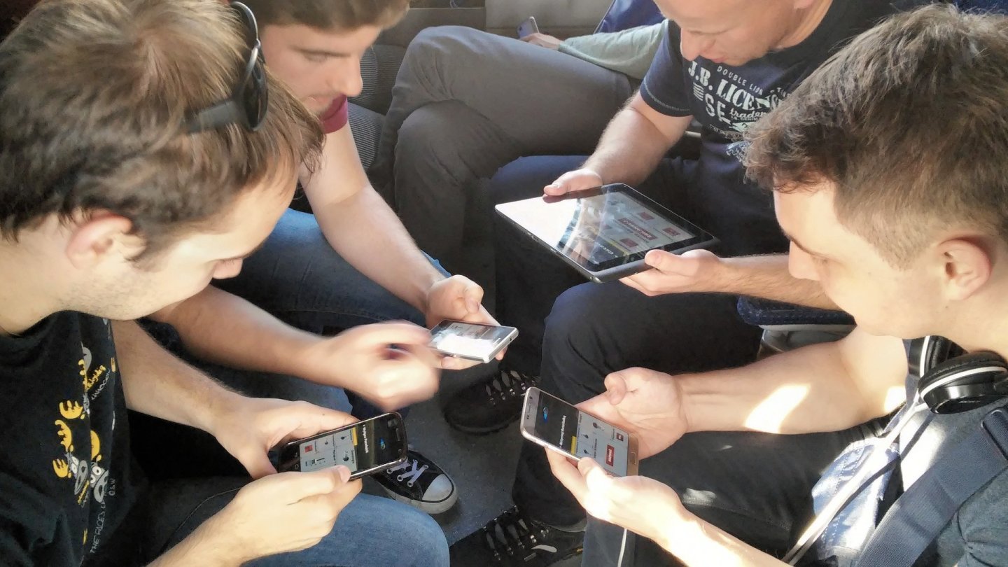 Patru tineri așezați aproape unul de altul, fiecare cu un telefon sau o tabletă în mână, jucând același joc multiplayer