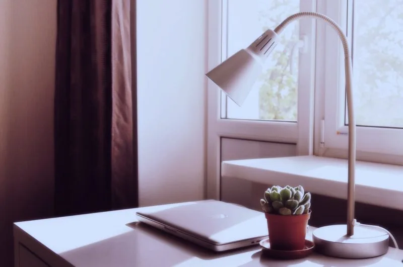 Laptop deschis pe o masă albă, lângă o lampă de birou și o plantă
