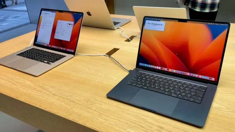 Două laptopuri MacBook Air deschise pe o masă de lemn, cu ecranele aprinse și imagini de fundal portocalii