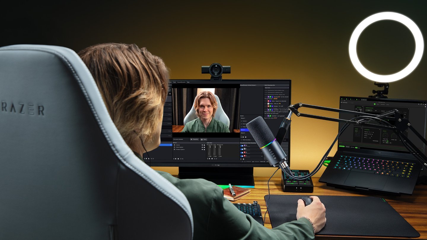 Creator de conținut așezat la birou în fața unui monitor cu camera Razer Kiyo V2 Pro deasupra, cu microfon pe braț, lampă inelară și laptop