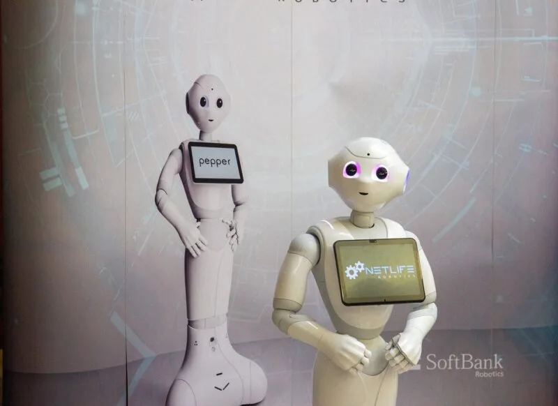 Doi roboți umanoizi Pepper cu tablete pe piept, expuși la un eveniment tehnologic