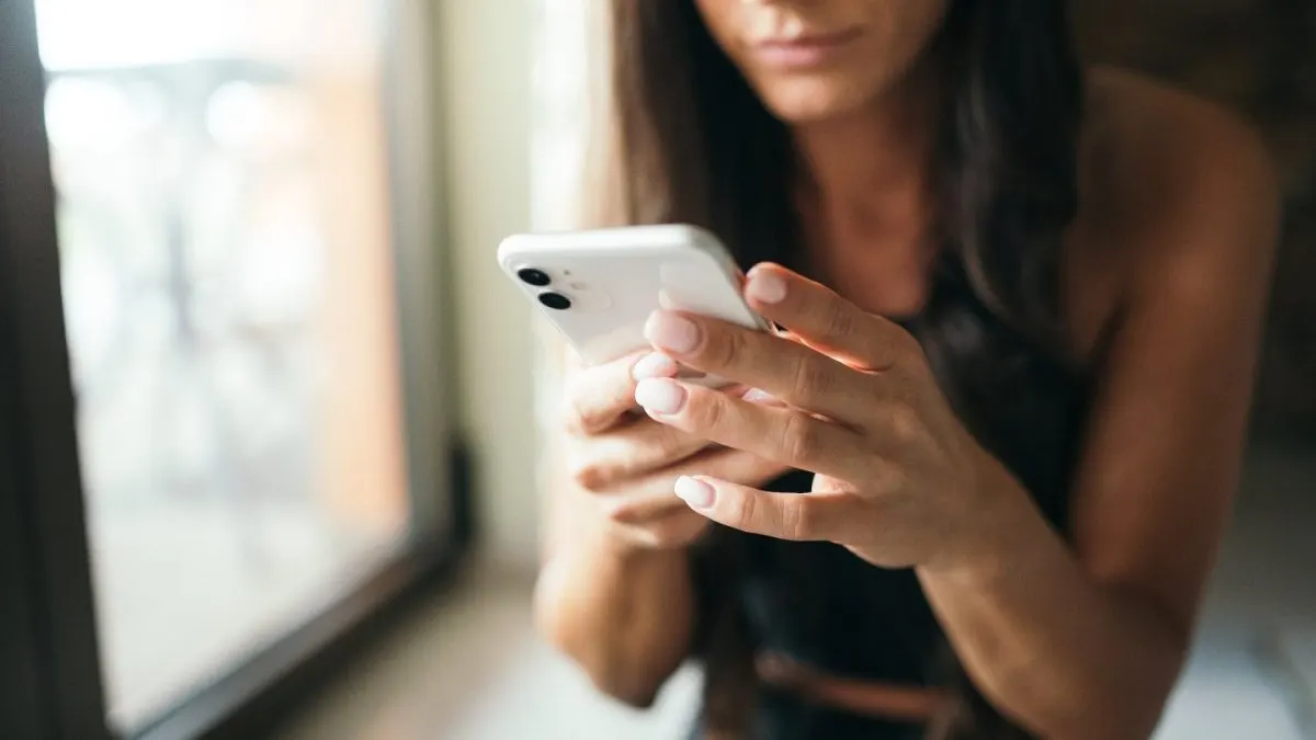 oferta YOXO Femeie care ține în ambele mâini un smartphone alb și tastează pe ecran, lângă o fereastră luminoasă