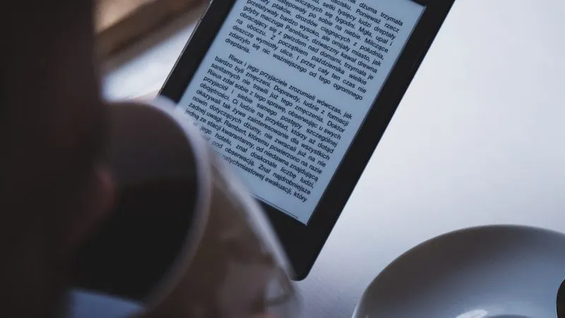 Persoană care citește pe un e-reader cu ecran e-ink, ținut în mână deasupra unei mese