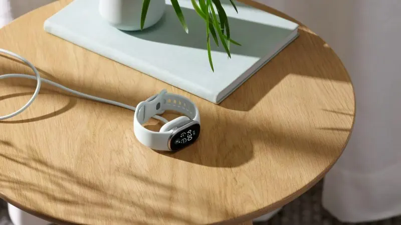 Brățara Google Fitbit Edge argintie, așezată pe o masă de lemn lângă încărcătorul magnetic