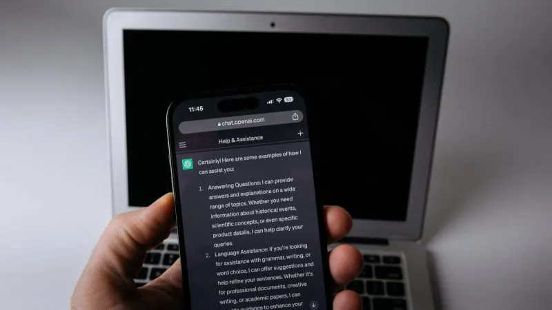 Mână care ține un smartphone cu o conversație ChatGPT deschisă, în fața unui laptop