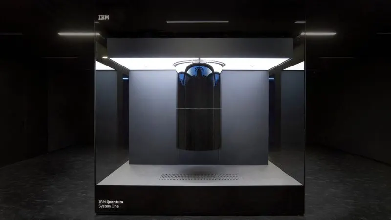 Calculatorul cuantic IBM Q System One, o carcasă de sticlă cu criostatul suspendat în interior, într-o sală întunecată