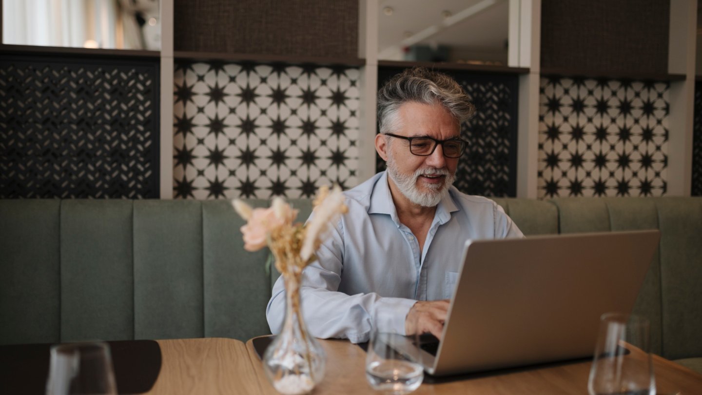 camera web a laptopului Bărbat cu ochelari care lucrează pe un laptop argintiu la o masă dintr-o cafenea, cu perete decorativ în spate