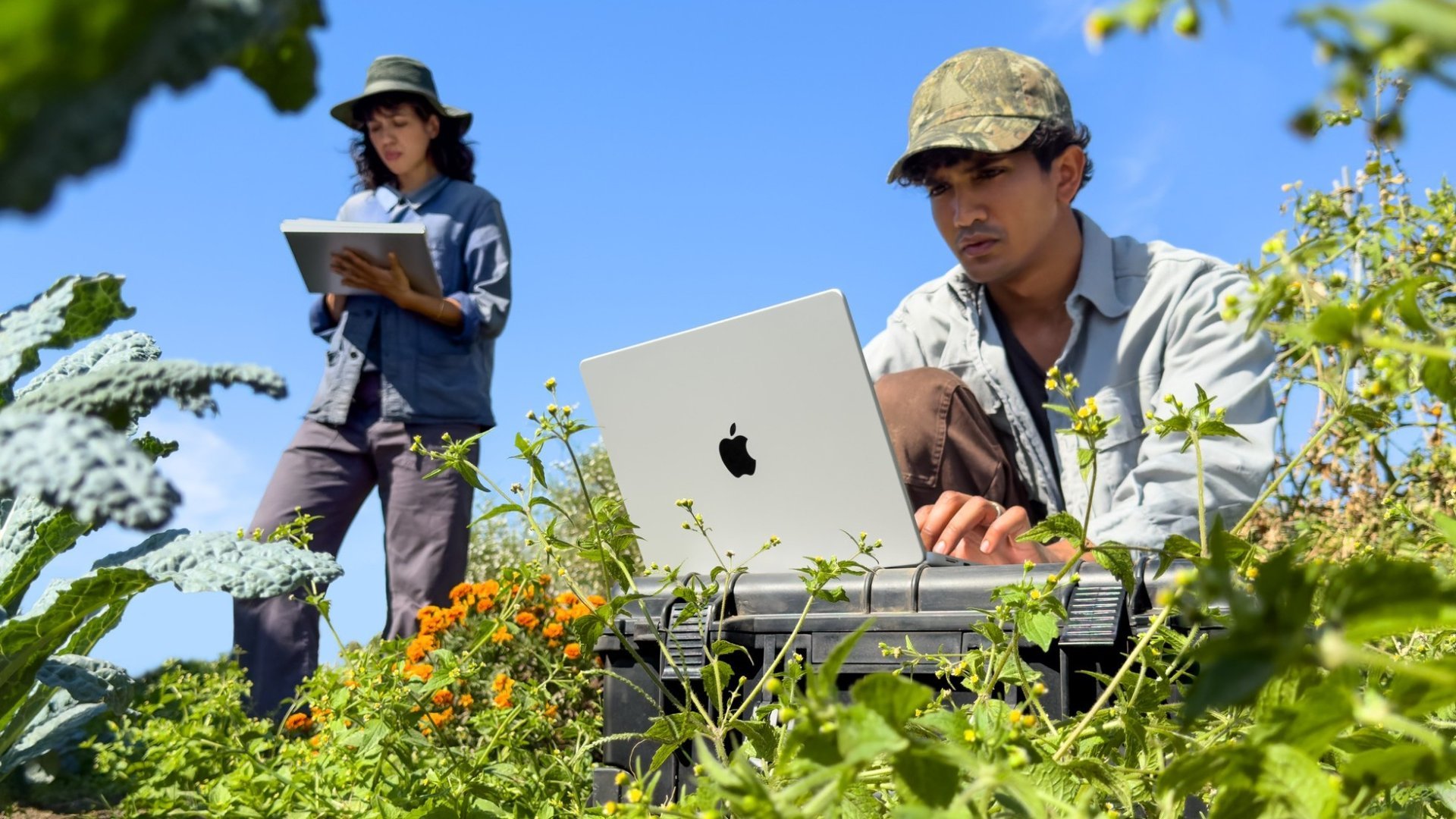 MacBook Pro cu M6 Doi cercetători lucrează pe teren, unul pe un MacBook Pro cu M6 argintiu, de 14 inchi, așezat pe o cutie, celălalt pe o tabletă