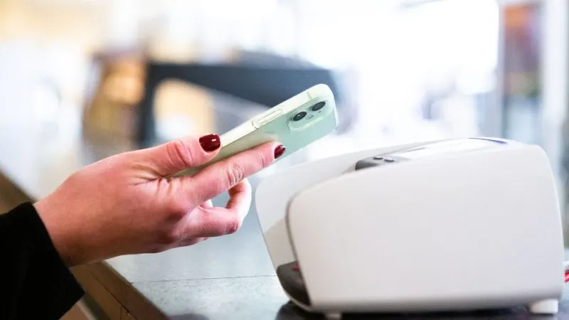 Mână care apropie un telefon mobil de un terminal de plată contactless, pe tejgheaua unui magazin