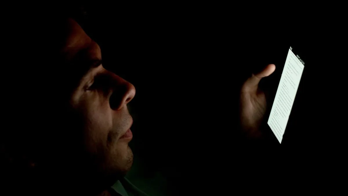 Reddit cere cont Bărbat care citește pe telefon într-o cameră întunecată, cu fața luminată de ecran