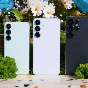Trei telefoane Samsung Galaxy S25 în culori diferite, așezate vertical printre flori și mușchi