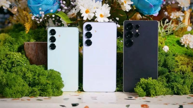 Trei telefoane Samsung Galaxy S25 în culori diferite, așezate vertical printre flori și mușchi