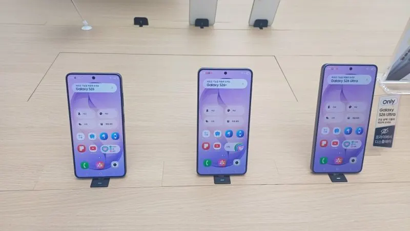 Trei telefoane Samsung Galaxy S26 așezate pe suporturi albe, cu ecranul principal pornit, într-un magazin