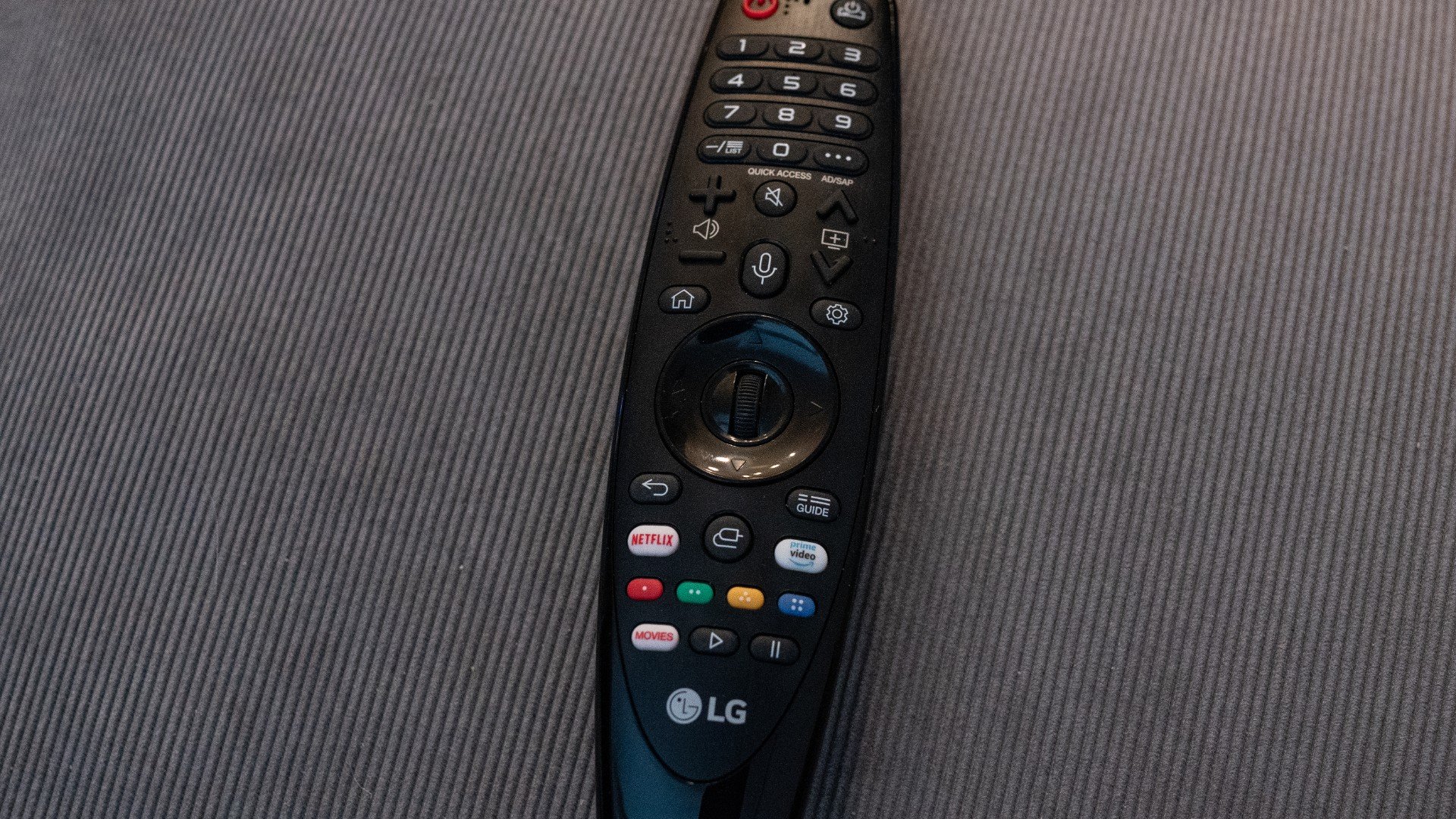Telecomandă LG pentru smart TV, cu butoane dedicate aplicațiilor de streaming, pe o canapea gri