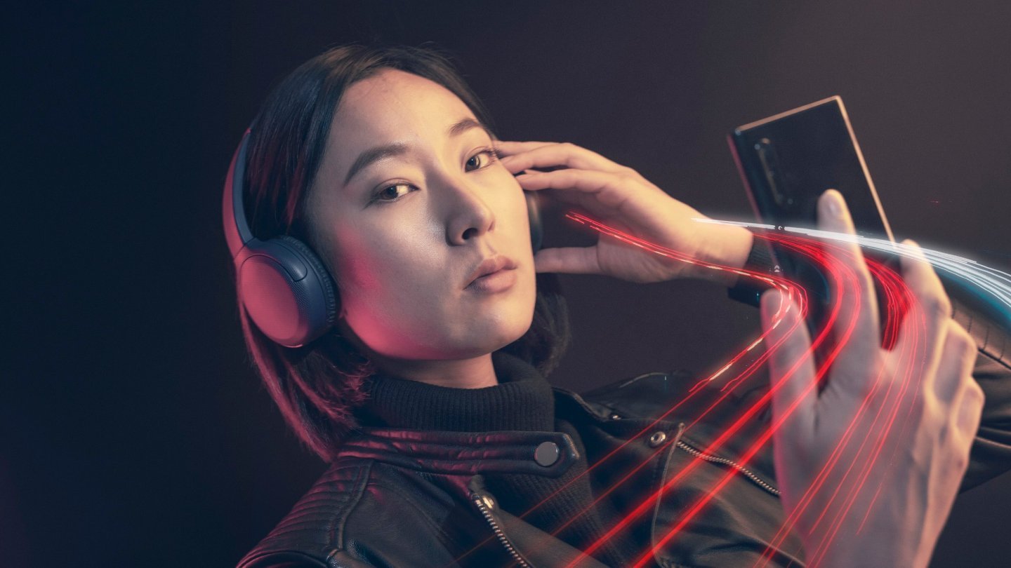 oferta YOXO Tânără cu căști over-ear și geacă de piele care privește un smartphone, pe fundal întunecat cu dâre de lumină roșie